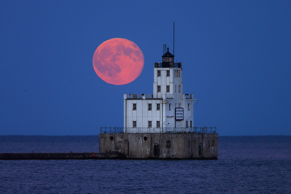 The blue moon, illuminated red by the setting sun, rises over Lake Michigan and the Milwaukee Breakwater Lighthouse.&nbsp;